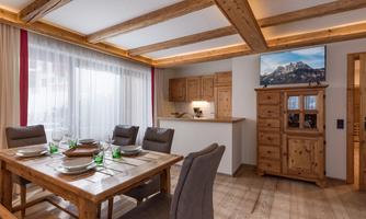 A cozy dining area with wood details and a table for six people. In the background, a modern kitchen and a television displaying a mountain landscape can be seen.
