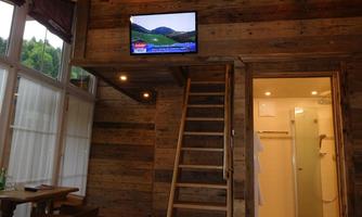 A cozy room with wooden walls and a television on the wall. A wooden staircase leads to an upper area, and an adjacent bathroom is visible.