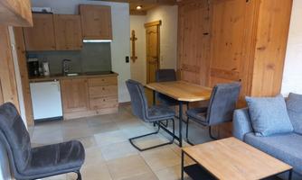 A cozy living kitchen with wooden paneling and modern furniture. The room includes a dining table with chairs, a small kitchen, and a comfortable sofa.