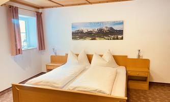 A large bedroom with a double bed and two pillows. On the wall hangs a picture of a mountain landscape, and there is a small table and a chair.