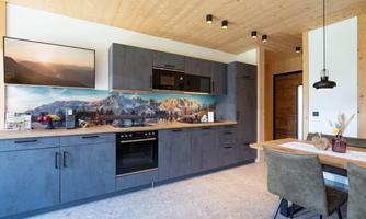 A modern kitchen with a wooden ceiling and stylish gray furniture. An impressive landscape photo is visible on the wall, and a dining table with chairs is located in the room.