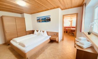 A cozy bedroom with a double bed and wood paneling. In the background, an adjacent dining area is visible.