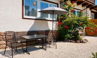 A beautiful outdoor seating area with a table and chairs, surrounded by blooming plants. A sunshade provides shade on a sunny day.