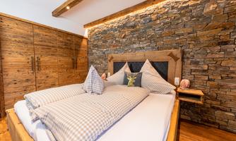 A cozy bedroom with a wooden bed and a stone wall accent. The bedding is in a checked pattern and there are wooden furniture in the room.