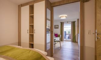 A modern furnished bedroom with a wardrobe and access to a living area. The room has plenty of daylight and a friendly atmosphere.