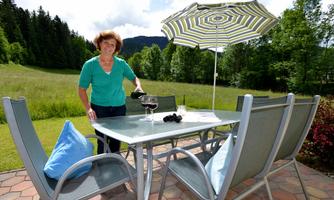 A woman is standing at a table outdoors, pouring wine. In the background, green meadows and trees can be seen.