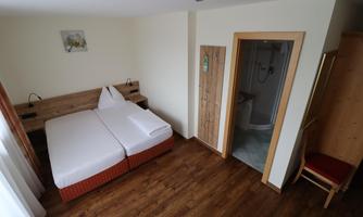 A simple room with two beds, a bedside table, and a chair. In the background, there is a bathroom with a shower.