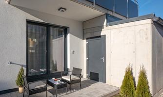 A cozy outdoor area with two chairs and a table. The wall is designed in a modern style, and there are small plants in the background.