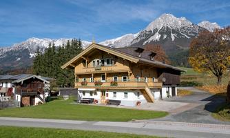 A traditional Alpine house with a wooden facade stands before an impressive mountain landscape. The surroundings are green and offer a picturesque view of the snow-capped peaks.