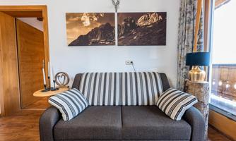 A cozy living room with a gray sofa and striped cushions. Two landscape pictures hang on the wall, and a window offers a view of nature.