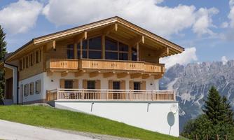 A modern wooden house with large windows and a balcony. In the background, majestic mountains and a blue sky are visible.