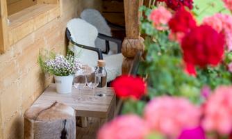 A cozy terrace with a small table, glasses, and a vase of flowers. Surrounded by colorful flowers and a warm wooden paneling.