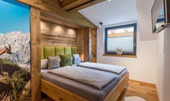 A cozy bedroom with a wooden bed and a large window. The walls are decorated with wood and a landscape graphic.