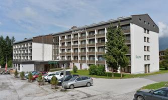 A modern hotel building in a quiet environment. Several cars and plants are in front of the hotel.