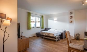 A modern living room with a sofa bed and a wooden table. Large windows with green curtains let in a lot of light.