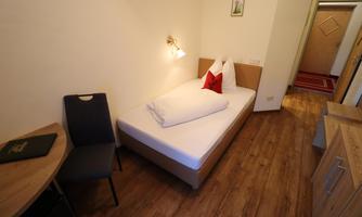 A simple room with a single bed, a chair, and a wooden desk. The walls are bright, and the room feels friendly and inviting.