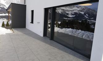 A modern house with large windows and a view of the snow-covered mountains. The floor is tiled with grey tiles.