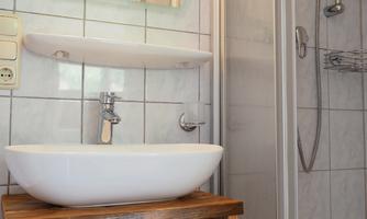 A modern bathroom with a sink on a wooden shelf. The shower is also in the room and one wall is tiled.
