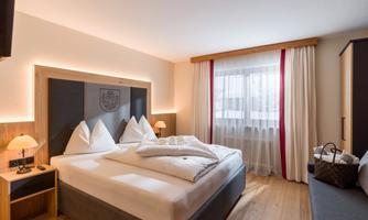 A modern hotel room with a large double bed and white pillows. The room has wooden flooring and large windows with bright curtains.