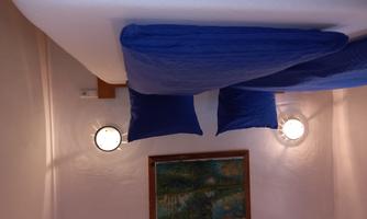 A cozy bedroom with two beds and blue cushions. A picture hangs on the wall and there are round wall lights.