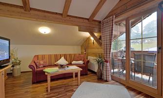 A cozy living room with wooden beams and a comfortable couch. The room has large windows overlooking a balcony and the surrounding nature.
