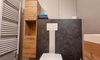 A modern bathroom with a white toilet and a wooden shelf. On the wall, a towel radiator and cabinets made of wood and gray material can be seen.