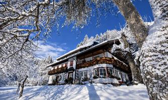 A picturesque winter house surrounded by snow-covered trees. The sky is clear and blue, creating a tranquil atmosphere.