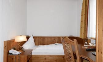A simple room with a single bed, a table, and a chair. There is also a lamp and a desk with a wooden appearance.