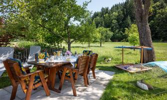 An inviting outdoor dining area with wooden furniture and blooming plants. In the background, a green meadow stretches out with trees.