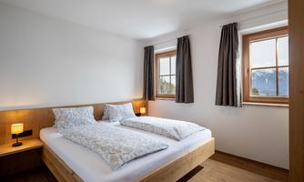 A bright bedroom with two beds and large windows. Wooden details and curtains create a cozy atmosphere.