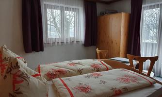 A cozy bedroom with a double bed and colorful pillows. Large windows let in plenty of light, and there is a wooden wardrobe and a chair in the room.
