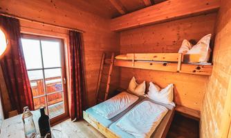 Dreibettzimmer Ein gemütliches Zimmer aus Holz mit zwei Betten, darunter ein Etagenbett. Große Fenster sorgen für viel Licht und bieten einen Blick nach draußen. 