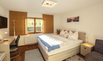 A bright bedroom with a large bed and cozy furnishings. A window features blinds and a plant.