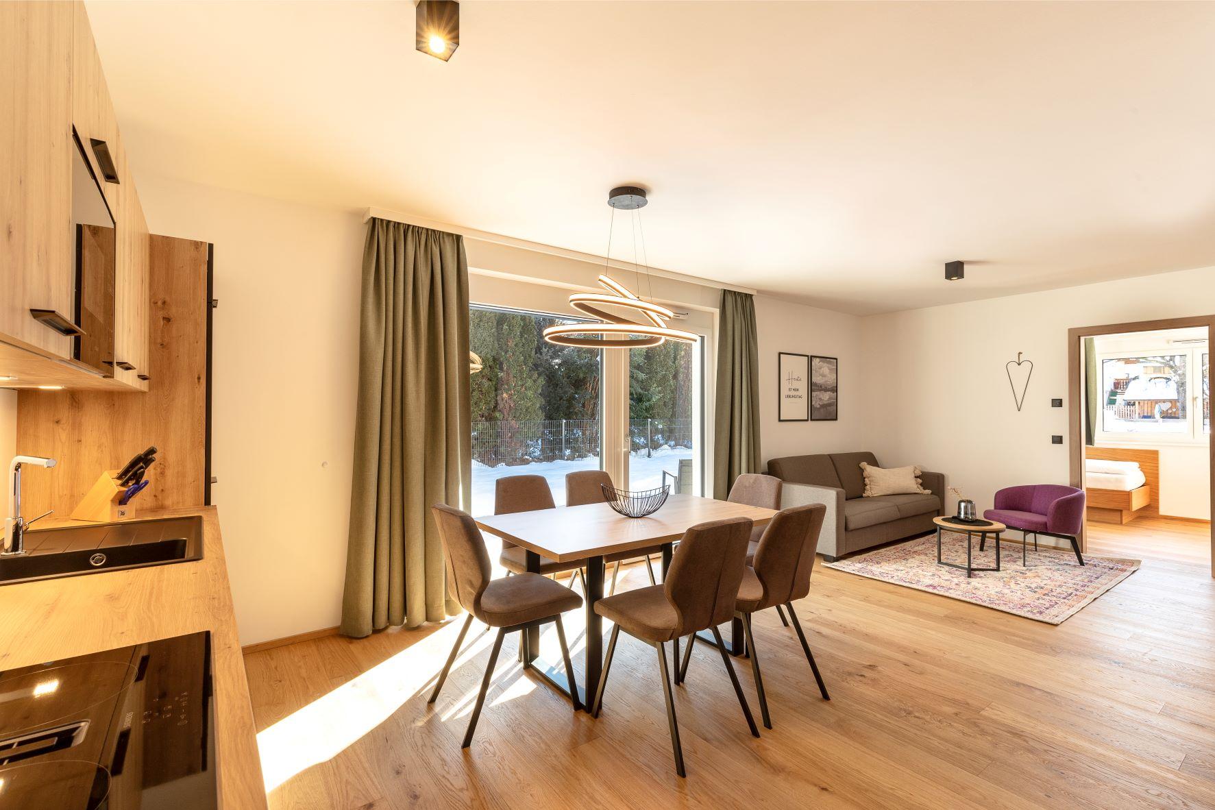 A modern living room with a dining table, chairs, and a cozy couch. Large windows let in plenty of daylight and offer a view outside.