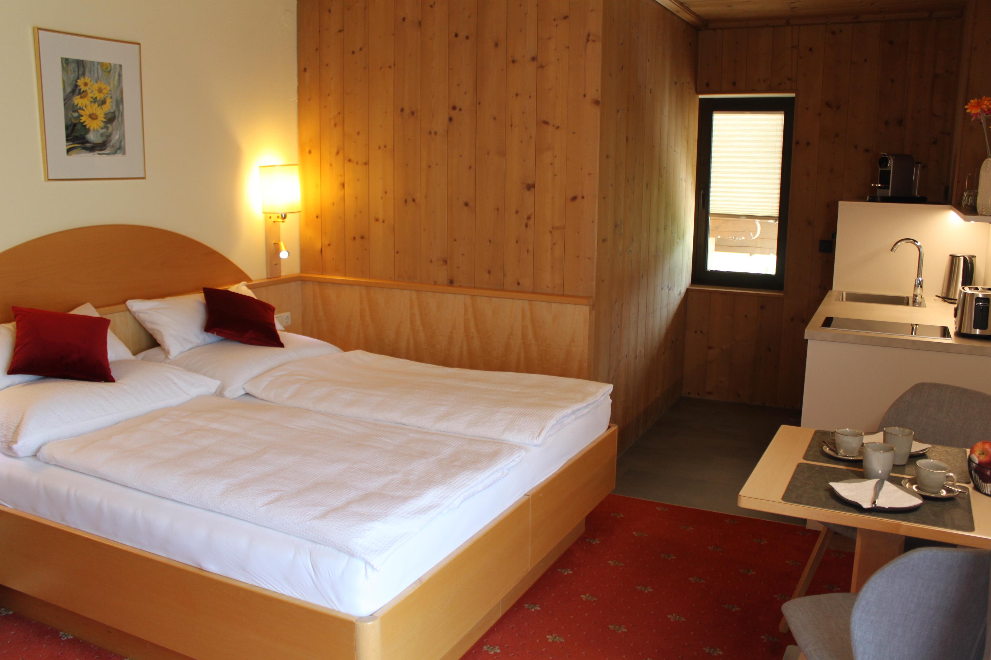 A cozy room with a double bed and a small kitchenette. The walls are decorated with wood, and there is a table for two.