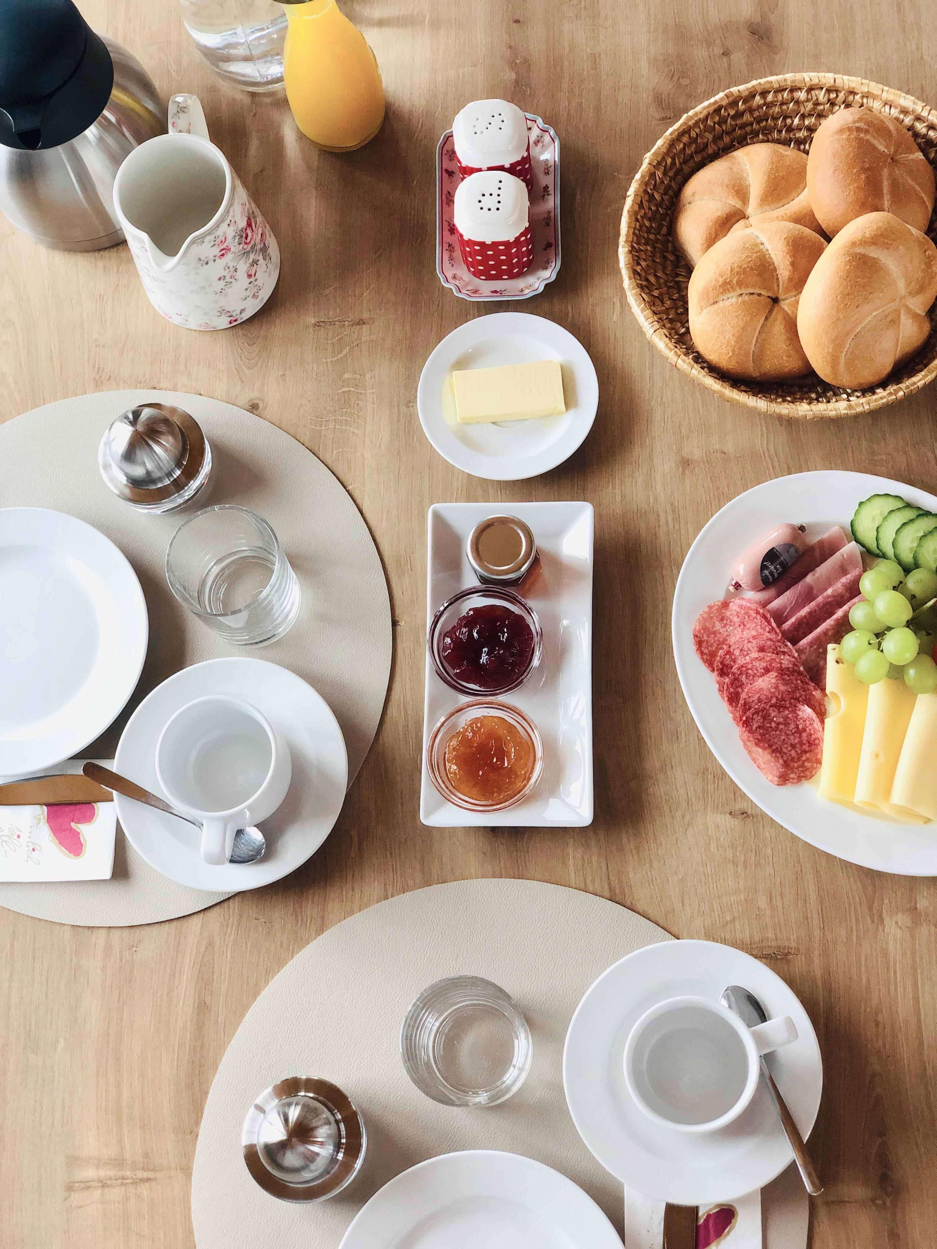 A beautiful breakfast table with rolls, sausage, cheese, and fresh fruit. There are also jam, butter, and drinks.