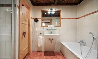 The stylishly furnished bathroom combines modern amenities with rustic charm. Features include a spacious bathtub with a handheld shower, an elegant sink with a glass shelf and large mirror, as well as a modern shower cubicle. A hairdryer and an adjustable cosmetic mirror complete the comfort. This bathroom offers a successful combination of functionality, comfort, and stylish design – ideal for relaxed moments.