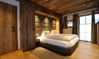 Spacious bedroom with modern chalet charm. This elegant bedroom impresses with its clean lines and high-quality furnishings. The comfortable double bed with a large, padded headboard is stylishly embedded in a reclaimed wood niche, providing a sense of coziness. The combination of natural materials, atmospheric lighting, and warm wooden floors creates a harmonious ambiance – ideal for relaxed nights and a restful stay in the alpine village.