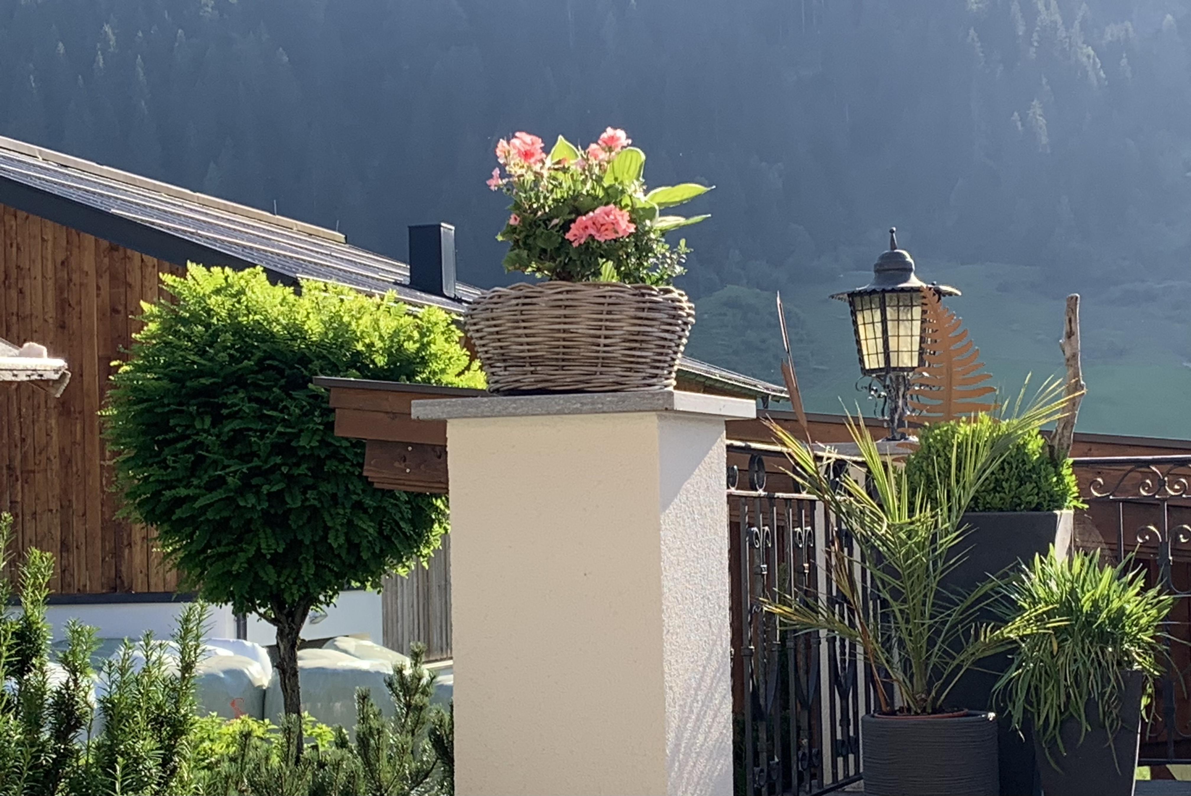 A terrace with a blooming plant pot and a pretty lantern. In the background, mountains and trees are visible.