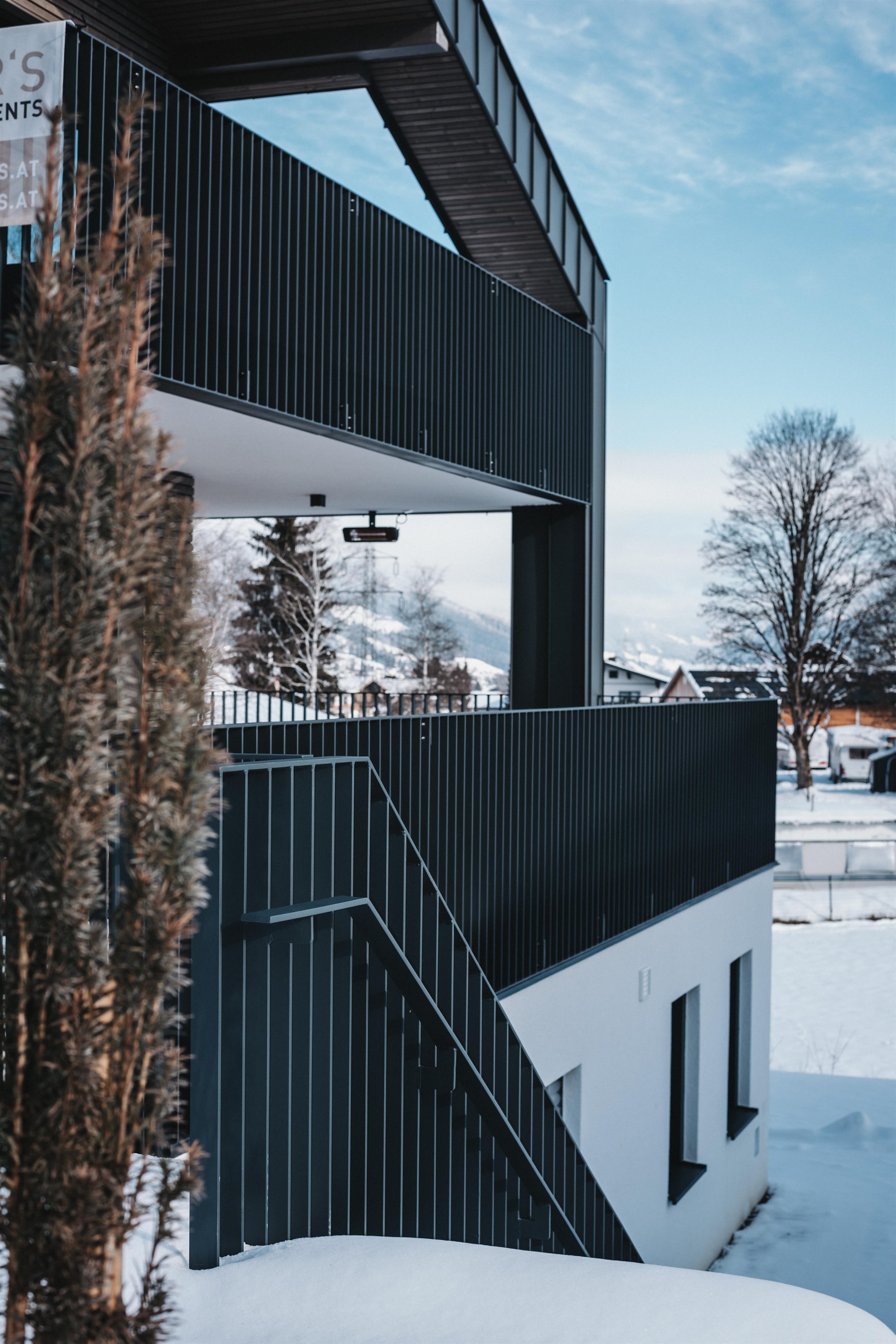 A modern building with dark facades and an outdoor staircase. Surrounded by snow and trees under a clear sky.