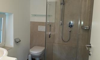 A modern bathroom with a glass shower and a white sink. The walls are in neutral tones, and a window provides daylight.