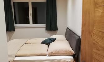 A cozy bedroom with a double bed and a wooden nightstand. The walls are light and there are curtains in dark green.