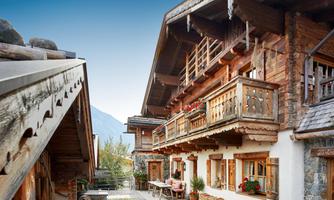 Authentic chalet architecture with attention to detail. This impressive exterior view showcases the chalet in all its beauty: traditional construction with natural stone, solid reclaimed wood, and artistically carved wooden balconies give the house its unique character. Flower boxes with red geraniums, shutters with heart motifs, and decoratively stacked firewood create an inviting and atmospheric ambiance. A cozy seating area in front of the house completes the harmonious overall picture – a place to arrive and feel at home.