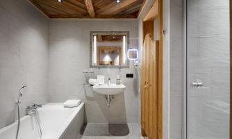 High-quality bathroom with alpine character. In this stylishly designed bathroom, modern comfort meets natural materials. Bright stone tiles, a spacious bathtub, as well as a shower and the elegant wooden ceiling create a calm, harmonious atmosphere. The integrated lighting concept at the mirror and high-quality fittings provide an exclusive feel-good experience – ideal for relaxing moments after an active day in the mountains.