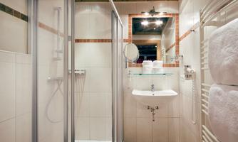 Modern bathroom with shower and all comforts<br/>This bright, well-maintained bathroom impresses with a modern shower with glass enclosure, a stylish washbasin, and a generous mirror with direct lighting. Warm tiles in natural tones and the visible wooden beams on the ceiling create a pleasant atmosphere. A cosmetic mirror, hairdryer, heated towel rail, and fresh towels complete the comfort offerings – ideal for a refreshing start to the day.