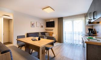 A modern living kitchen with a dining area and a cozy seating corner. Bright walls and large windows ensure plenty of daylight. A television is installed in the living bedroom.