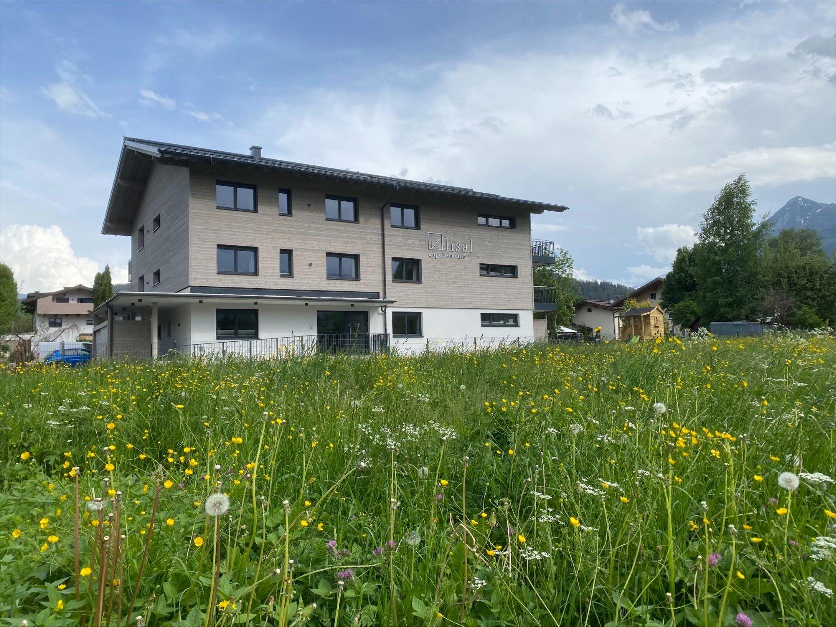 A modern building stands in the middle of a blooming meadow. In the background, trees and mountains can be seen.