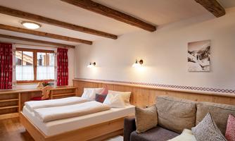 Cozy bedroom with double bed and sofa – relaxation in alpine style<br/>This bright and friendly bedroom combines Alpine charm with modern comfort. The spacious double bed with high-quality bedding invites restful nights, while the cozy sofa offers additional space to relax. Thoughtful details such as wooden beams on the ceiling, traditional heart curtains, and natural materials create a harmonious atmosphere. The integrated desk area with a view out the window makes the room ideal for quiet hours – whether for reading, working, or simply enjoying.
