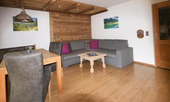 A cozy living room with a gray sofa and a wooden table. Pictures of landscapes hang on the walls, and the floor is made of wood.