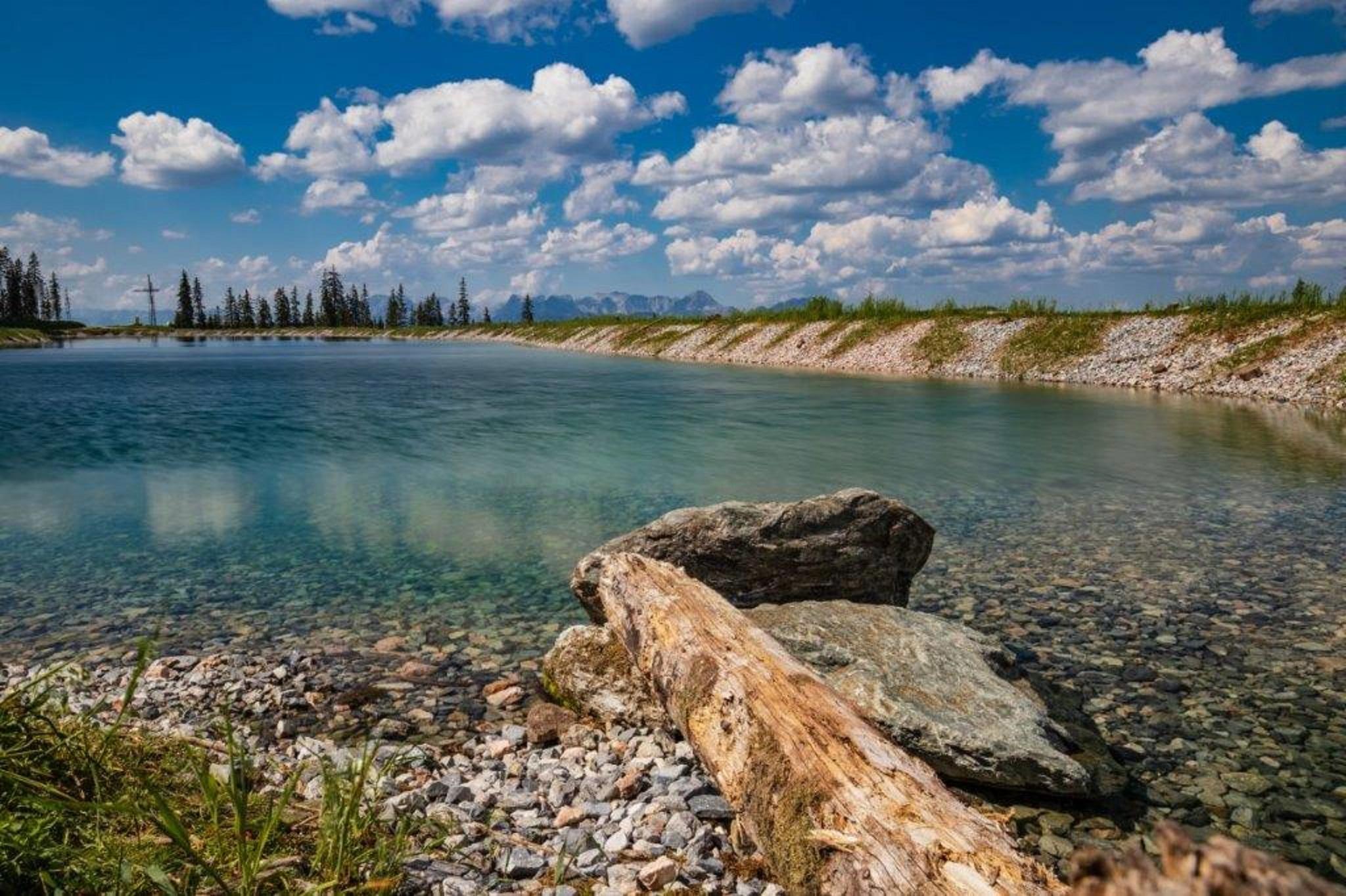 A calm lake with clear water and scattered stones. In the background are green meadows and lively clouds in the sky.